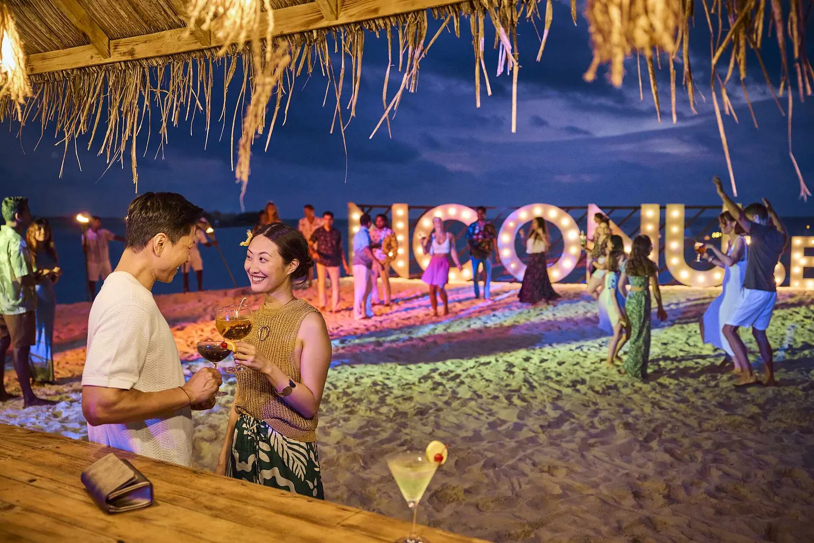 A night party on the beach, with coloured lights, the NOONU sign and guests dancing with cocktails in hand.