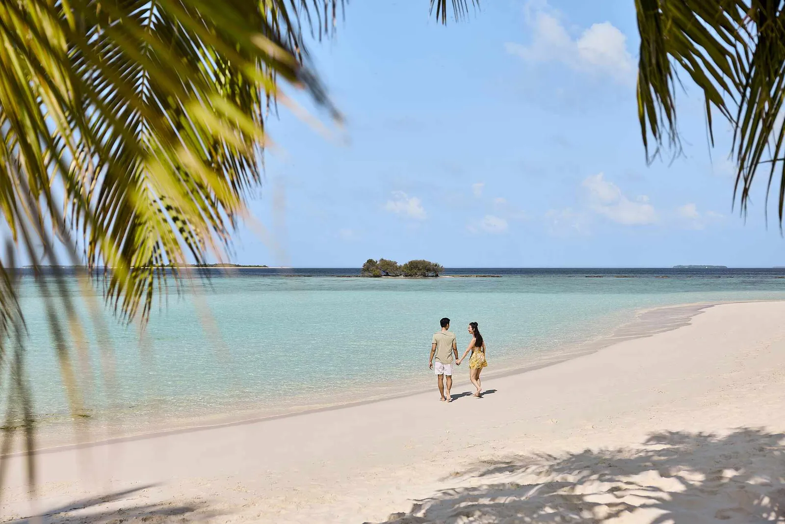 A couple walking along the water’s edge between the palms and the shallow lagoon, with a small island on the horizon.