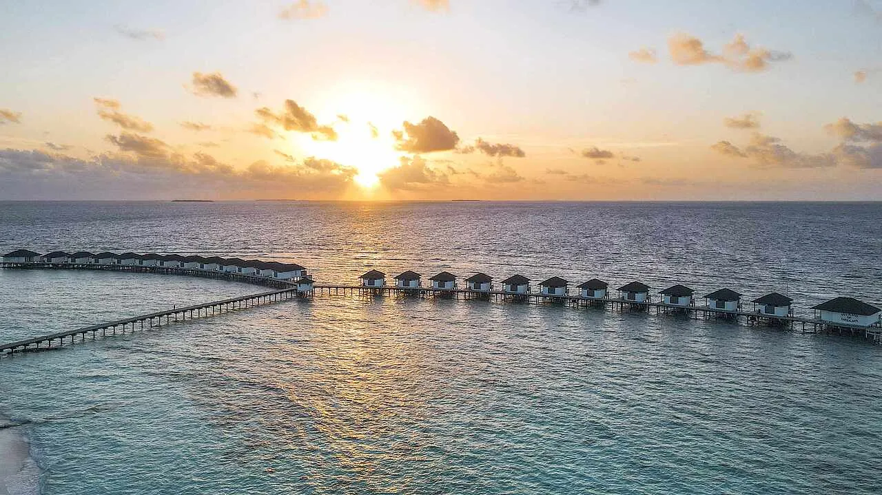 The sun setting over a flat sea behind the row of water villas, strung along their wooden jetty.