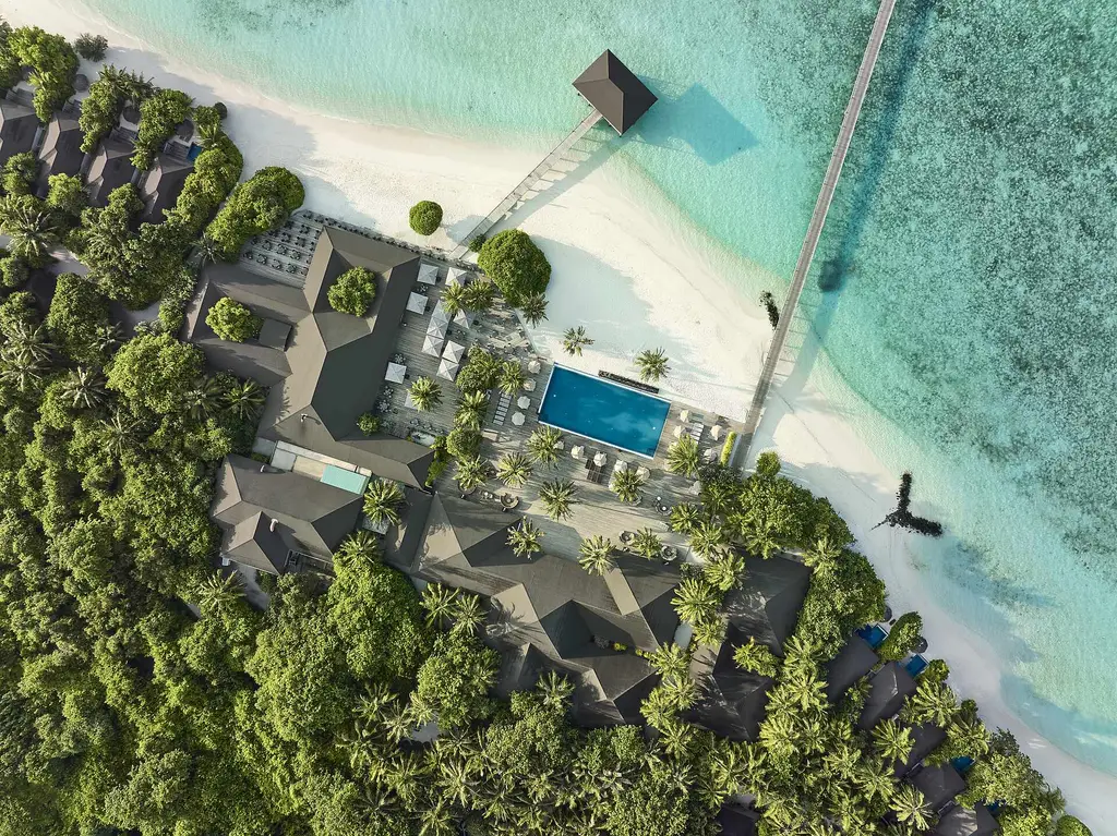 The heart of the island from above: dark roofs among the trees, the pool, the beach and the jetty out into the lagoon.
