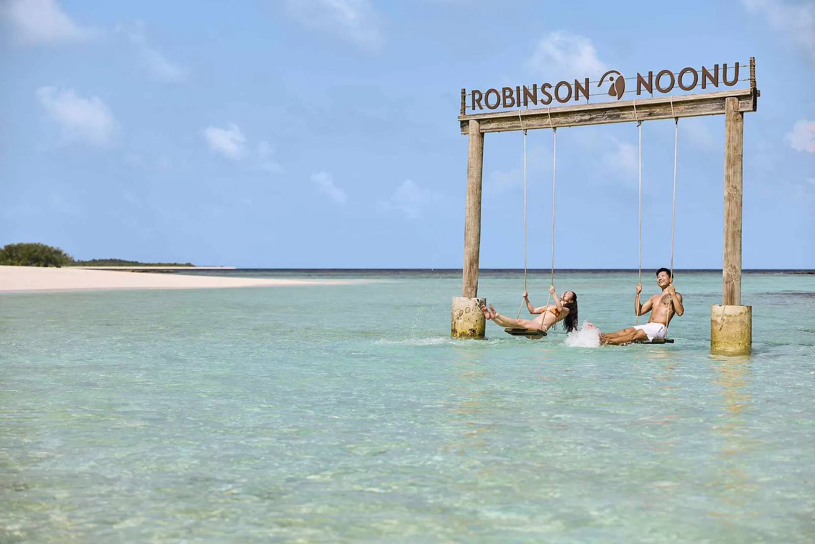 2 wooden swings under a ROBINSON NOONU sign set in the shallow lagoon, with a couple sitting over the water.