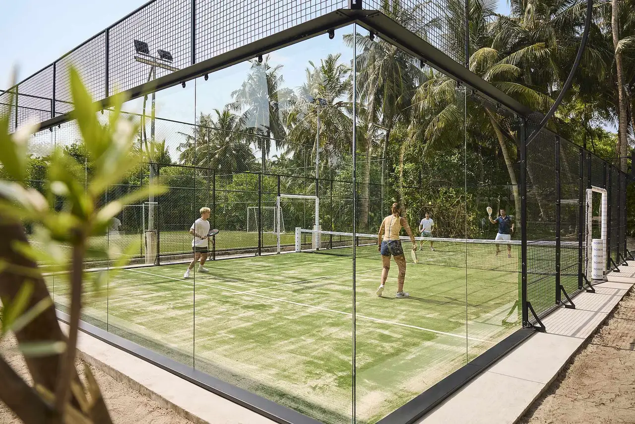 The glass-walled padel court among the palms, with 4 players mid-rally.