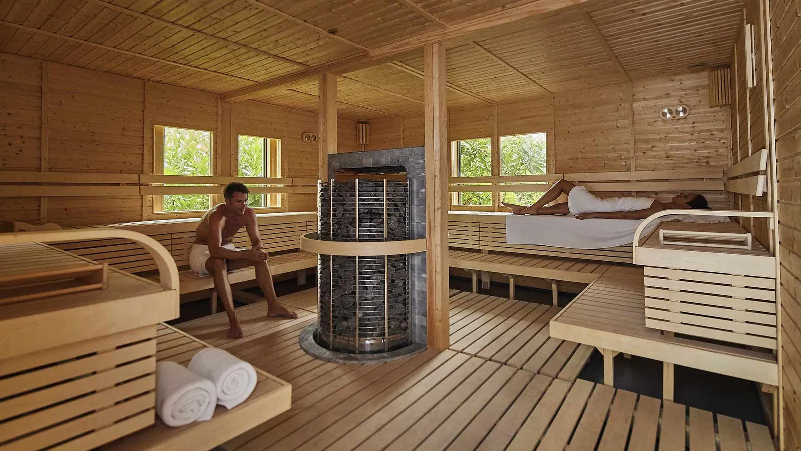 The Finnish sauna lined in pale wood, with the stove in the middle and 2 guests stretched out on the benches.