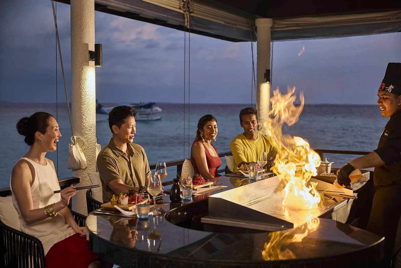 The teppanyaki chef raises a flame on the griddle in front of 4 guests, with the sea beyond the windows.