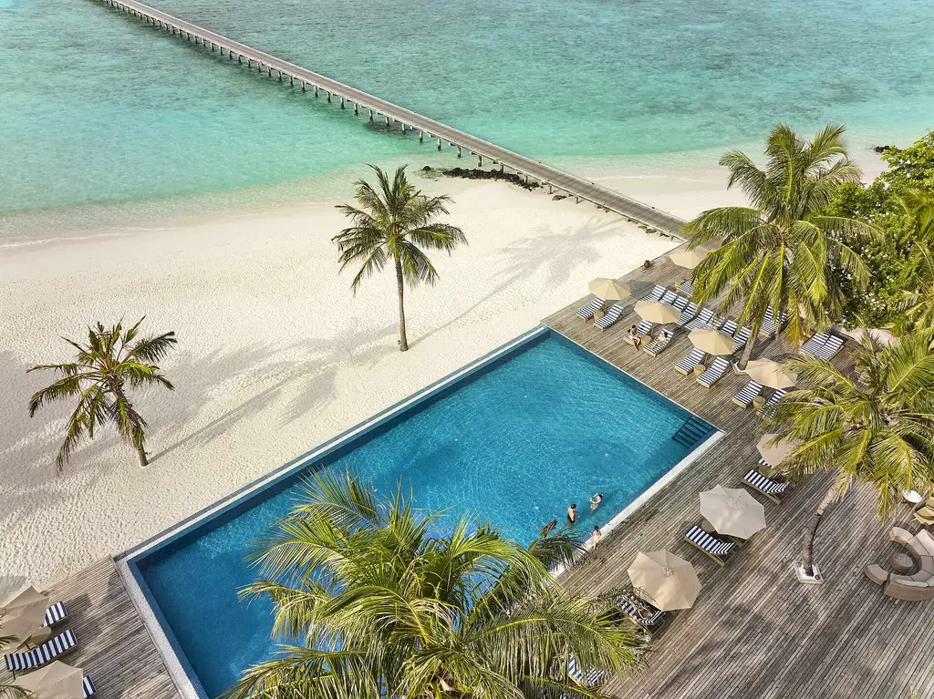 The infinity pool from above, with loungers on the timber deck, palms, the white beach and the long jetty over the lagoon.