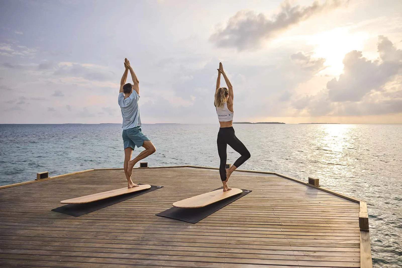 2 people balancing on yoga boards on the timber deck facing the sea at sunset.