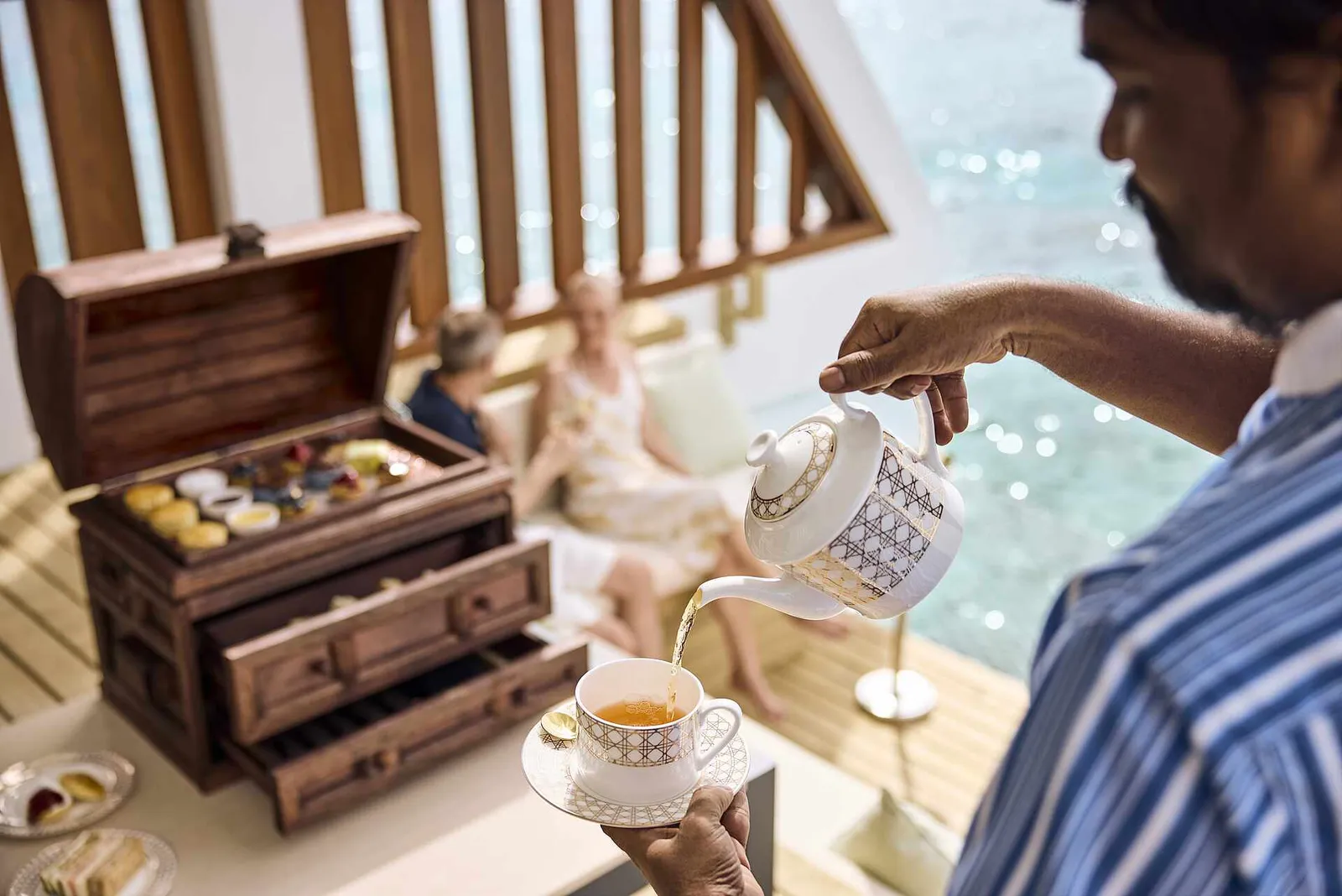 A waiter pours tea into a cup beside a wooden chest of small cakes, on a terrace above the sea.