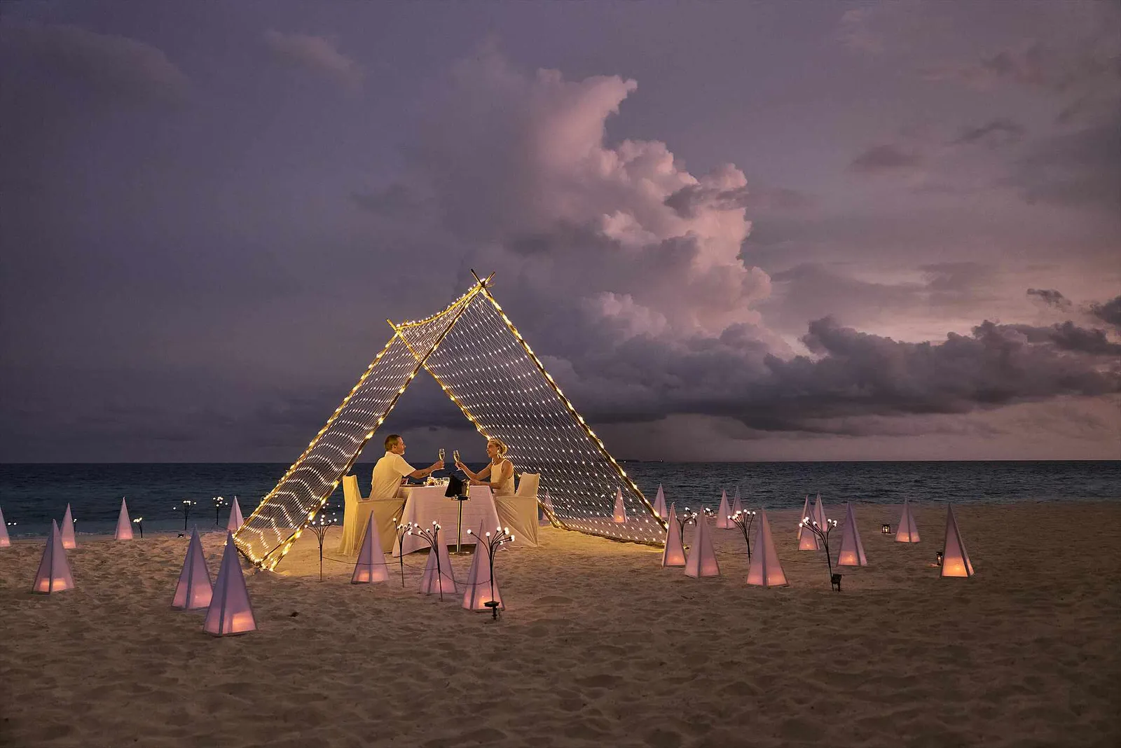 Dinner for 2 on the sand at dusk, under a tent of lights, among pyramid lanterns.