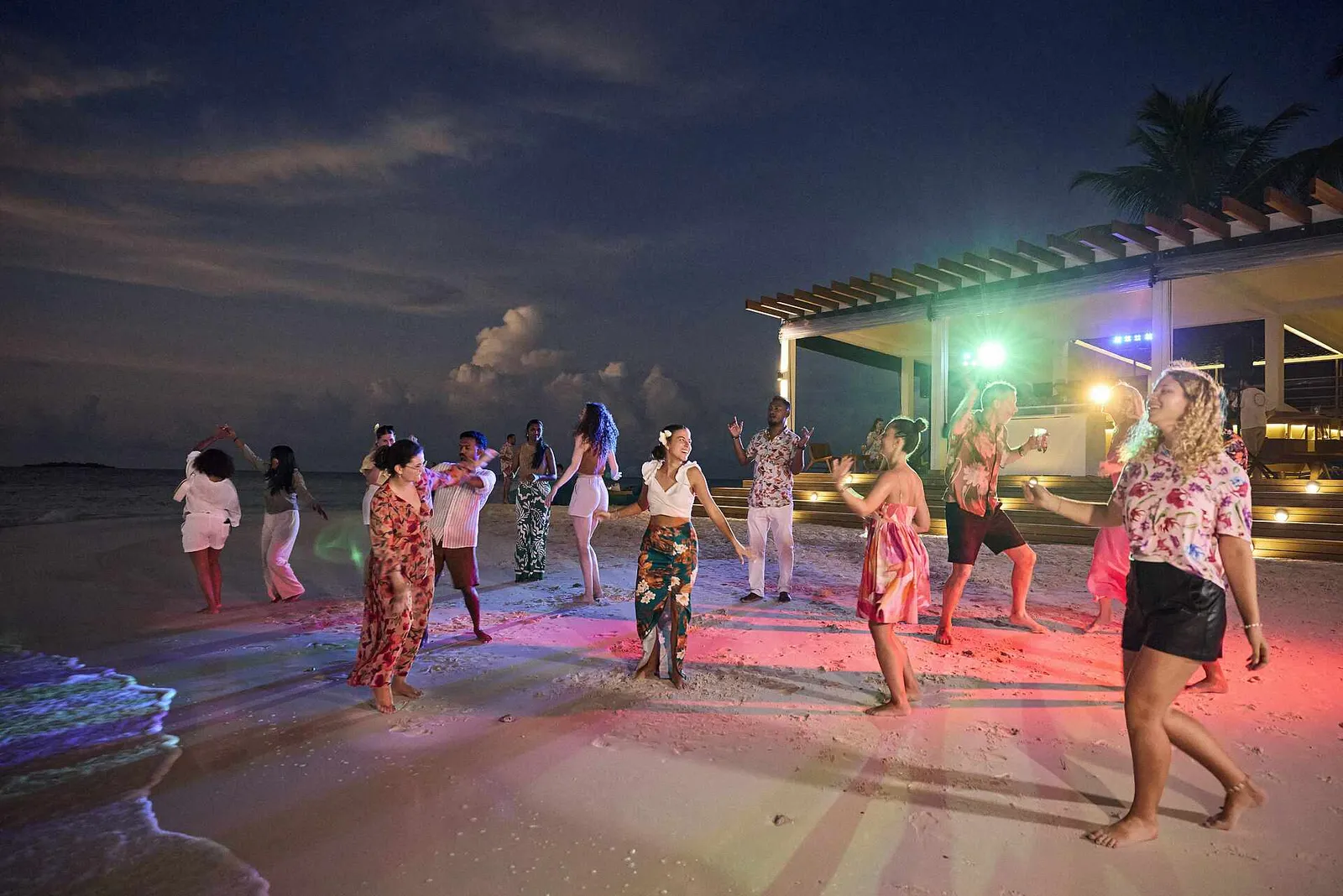 A night party on the beach, with coloured lights on the sand and guests dancing in front of the bar.