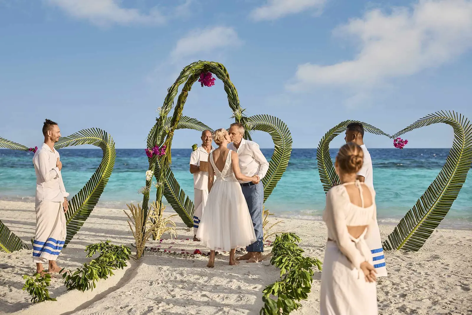 A ceremony on the beach: the couple under an arch of palm leaves and flowers, with the turquoise sea behind.
