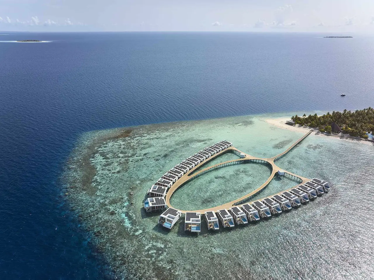 The ring of water villas from above, on the edge of the reef, with the island behind and open sea all round.