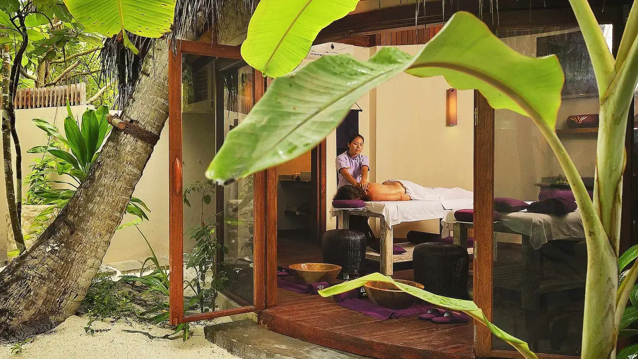 A spa room among the banana plants: the glass door open, a massage under way and a couple’s bed beside it.