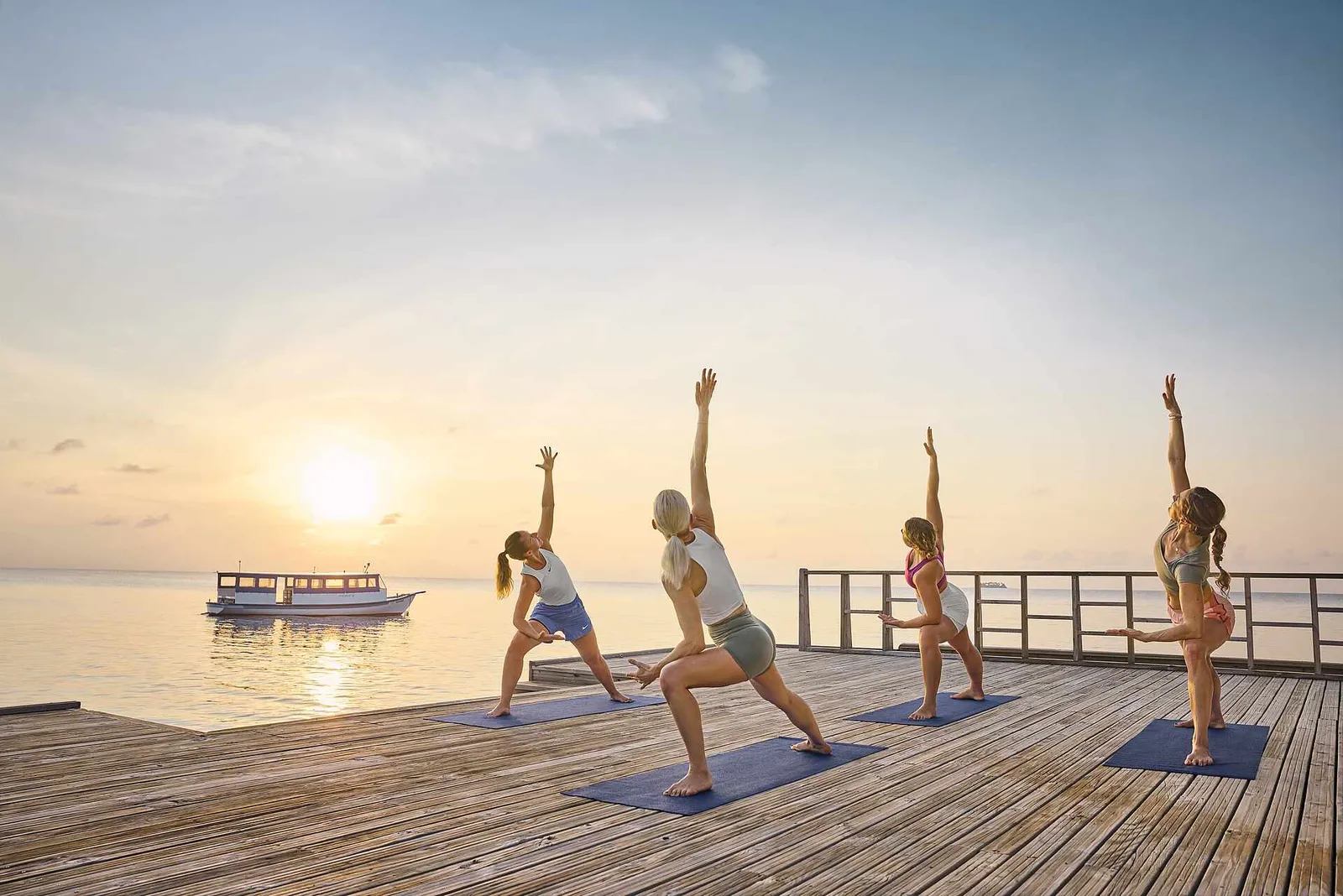 4 people doing yoga at sunrise on a timber deck over the sea, with a boat on the horizon.