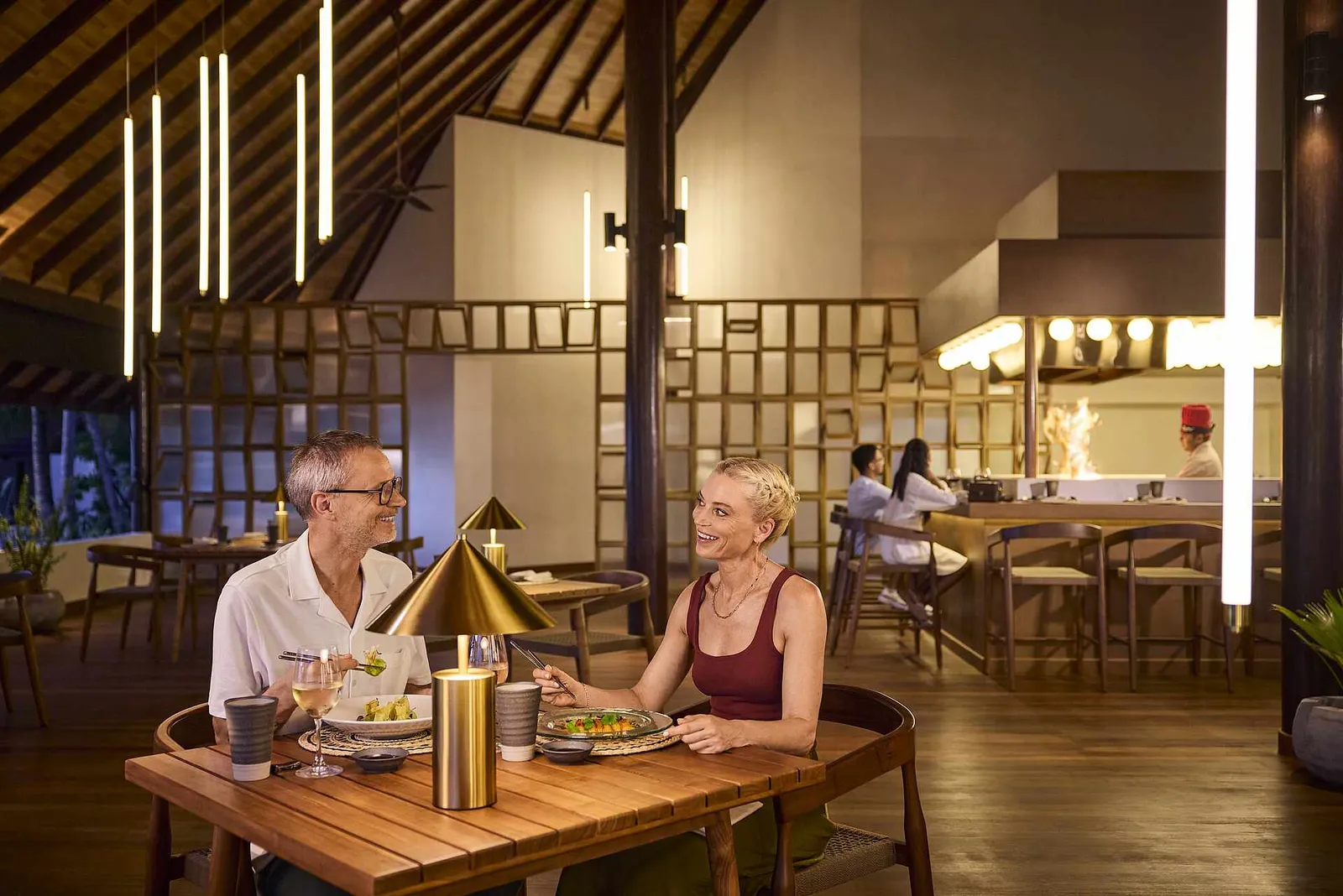 A couple at their table in the teppanyaki restaurant, under the timber ceiling, while the chef fires the griddle behind them.