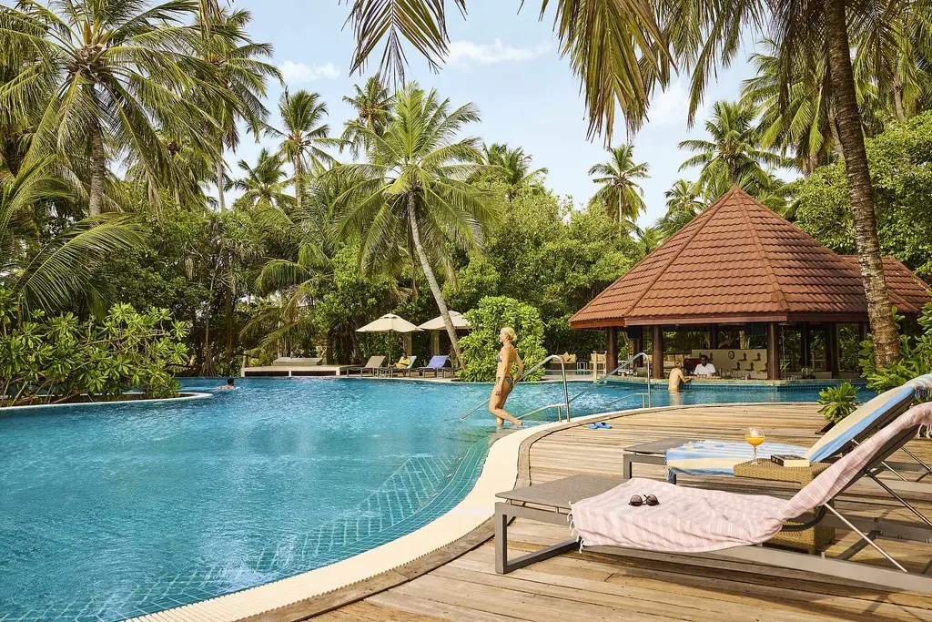 The main pool among the palms, with the red-roofed bar, loungers on the timber deck and a woman stepping into the water.