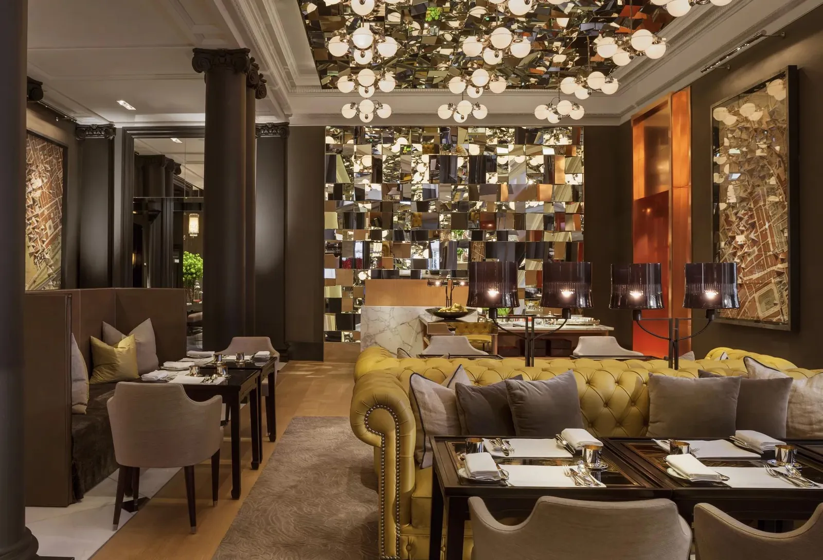 La Mirror Room vuota: il divano chesterfield giallo senape con i cuscini grigi, i tavoli apparecchiati, il soffitto e la parete di specchi, i lampadari a globi.