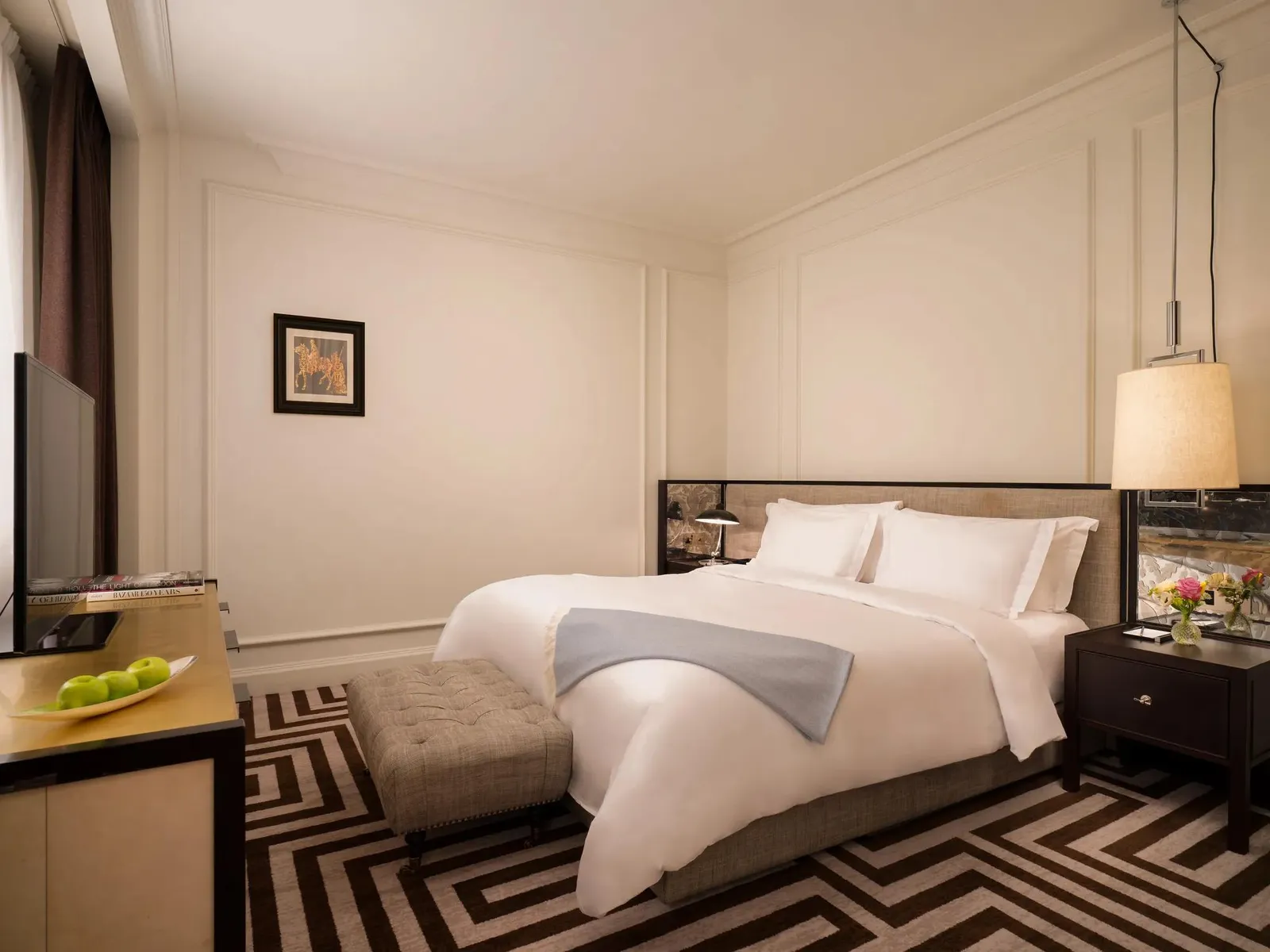 A Deluxe Room: a white bed with a pale blue throw, an upholstered bench, a black-and-white key-pattern rug and cream panelled walls