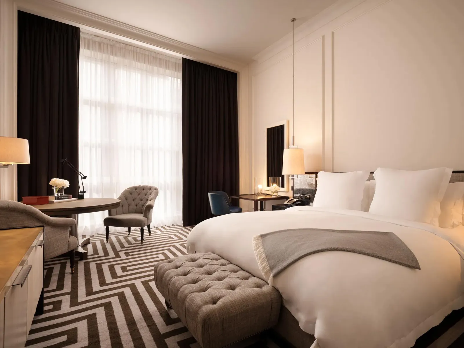 A Grand Premier Room: a white bed, a desk and armchairs beside 2 tall windows with dark curtains