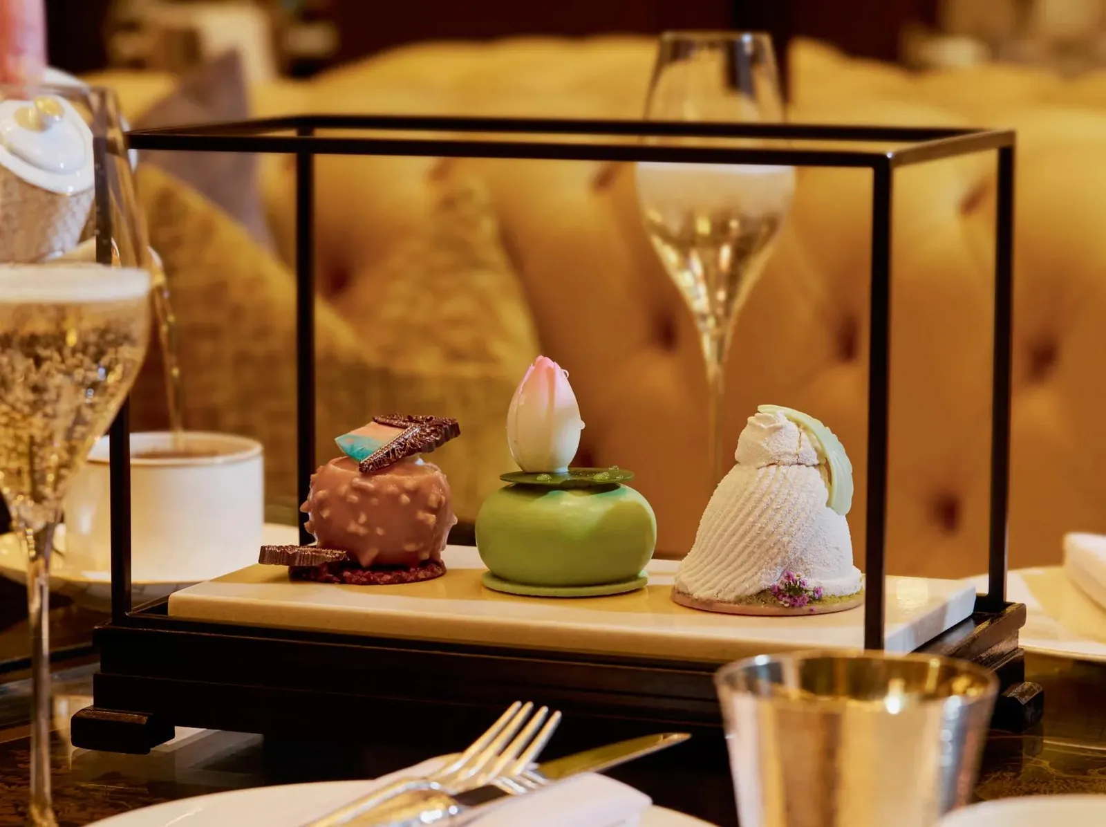 3 Art Afternoon Tea pastries beneath a glass-and-brass case, with a glass of Champagne beside them