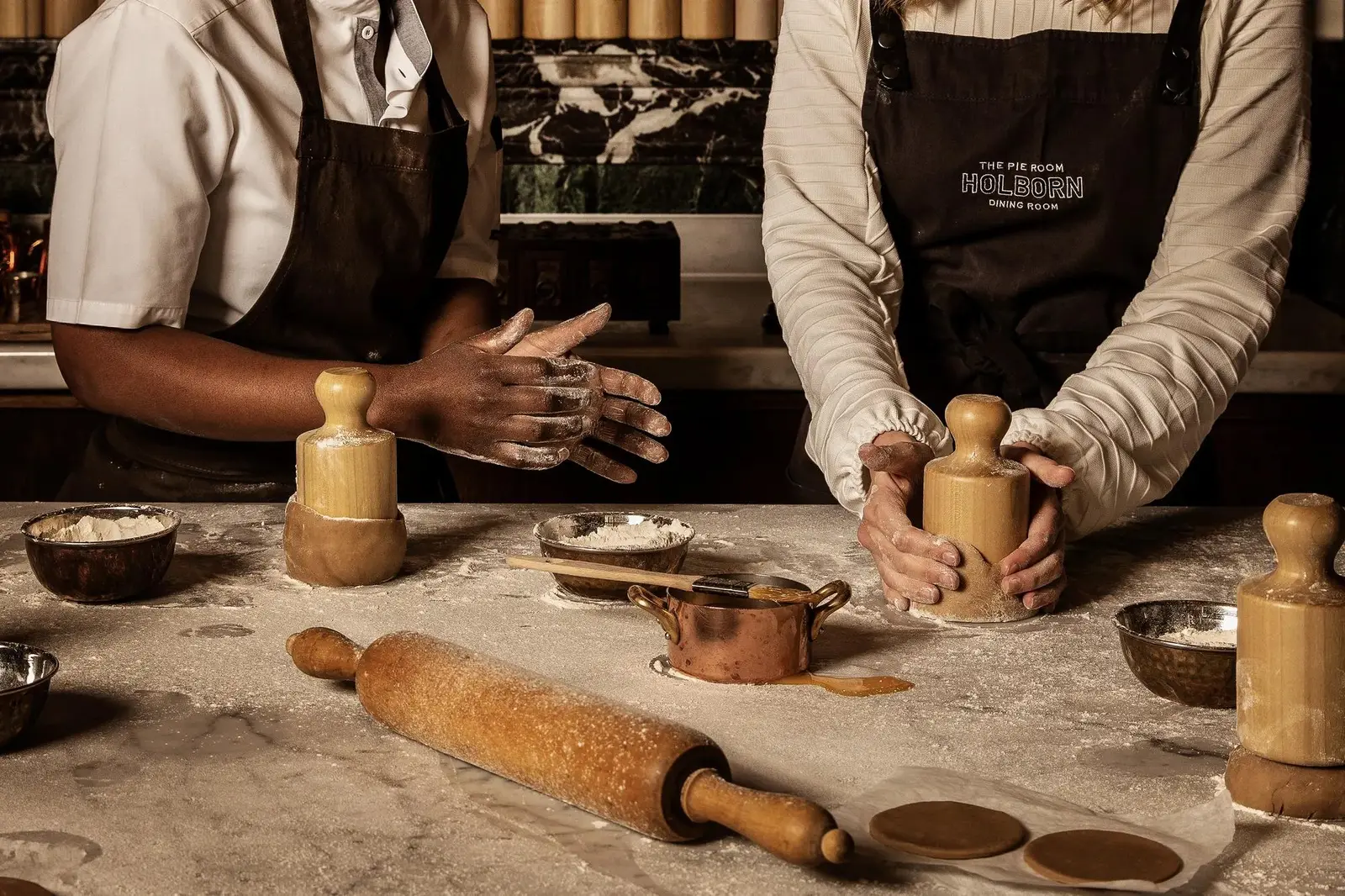 2 chefs in aprons work pie pastry on a marble counter, with a rolling pin and copper bowls