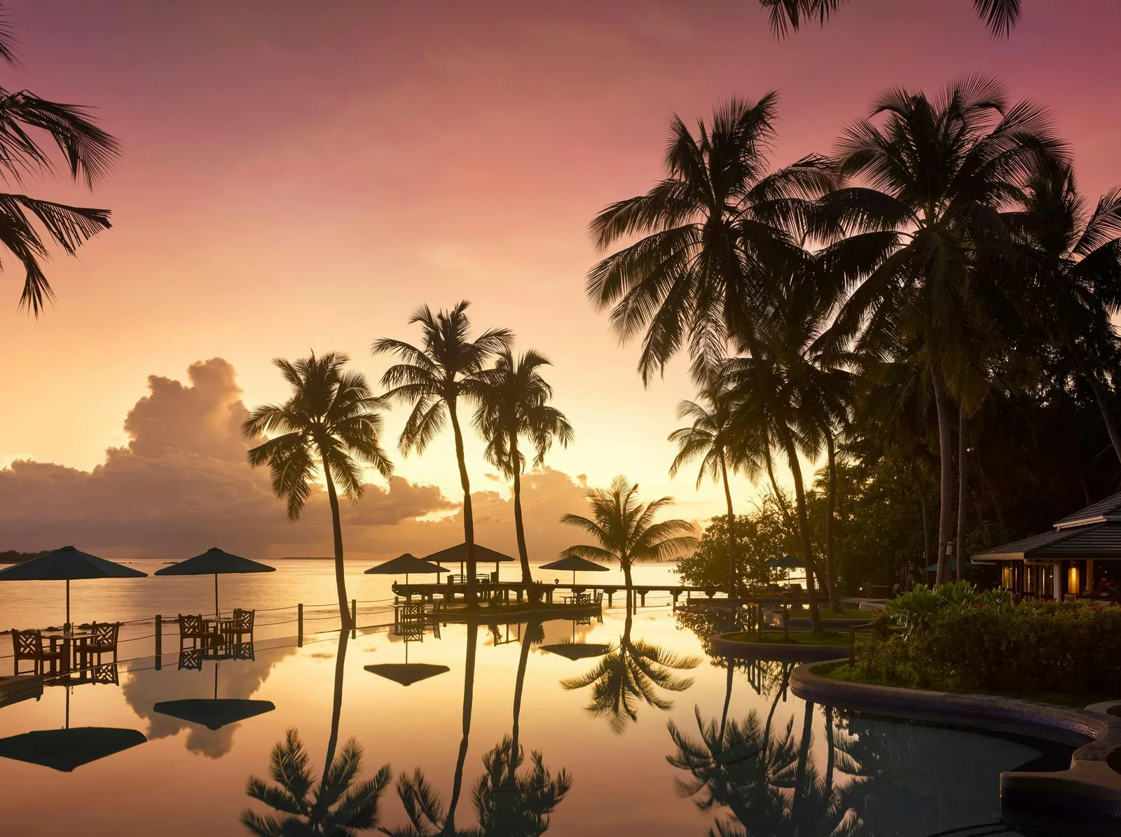 The main pool at sunset, palms and parasols reflected in the still water and the sea beyond.