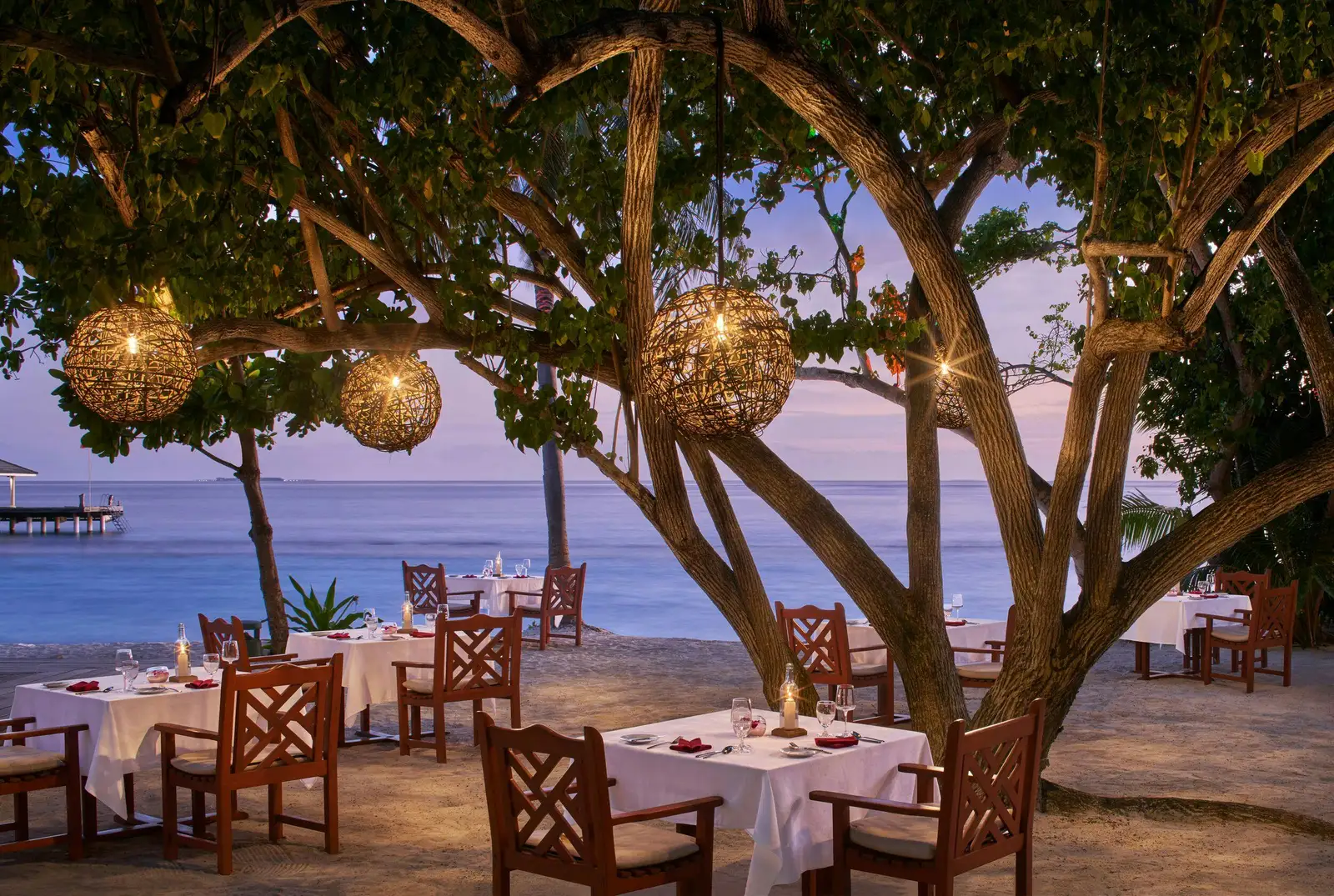 Raabondhi’s tables under a tree hung with rattan lanterns at dusk, the jetty out over the sea.