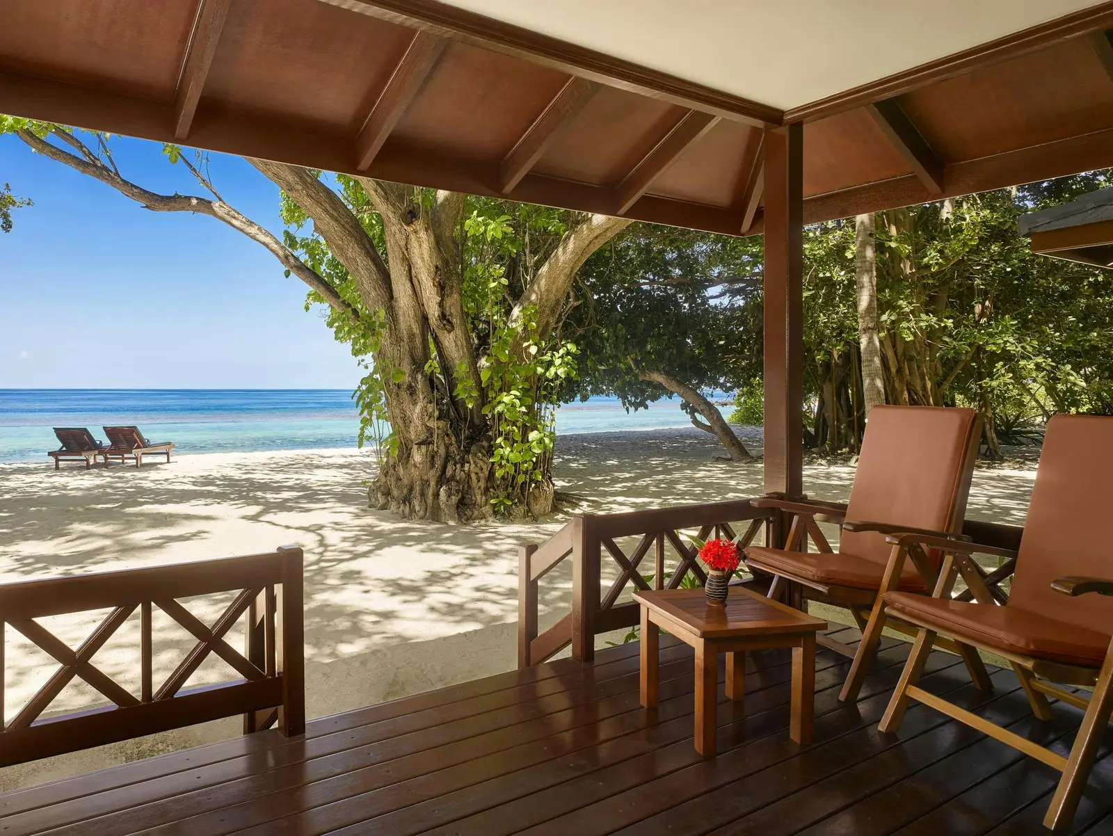 A Beach Villa’s timber veranda, 2 chairs and a small table, the big tree and the beach in front of the sea.