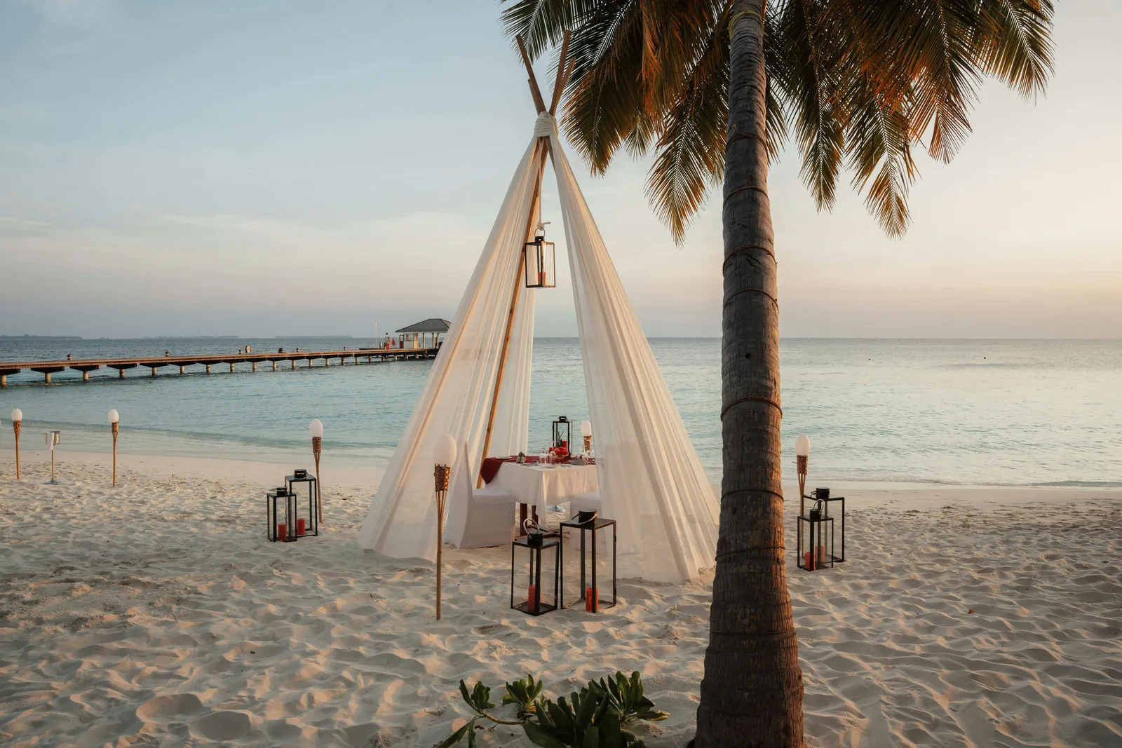 A table for 2 under a canvas canopy on the beach, among lanterns, with the jetty in the distance.