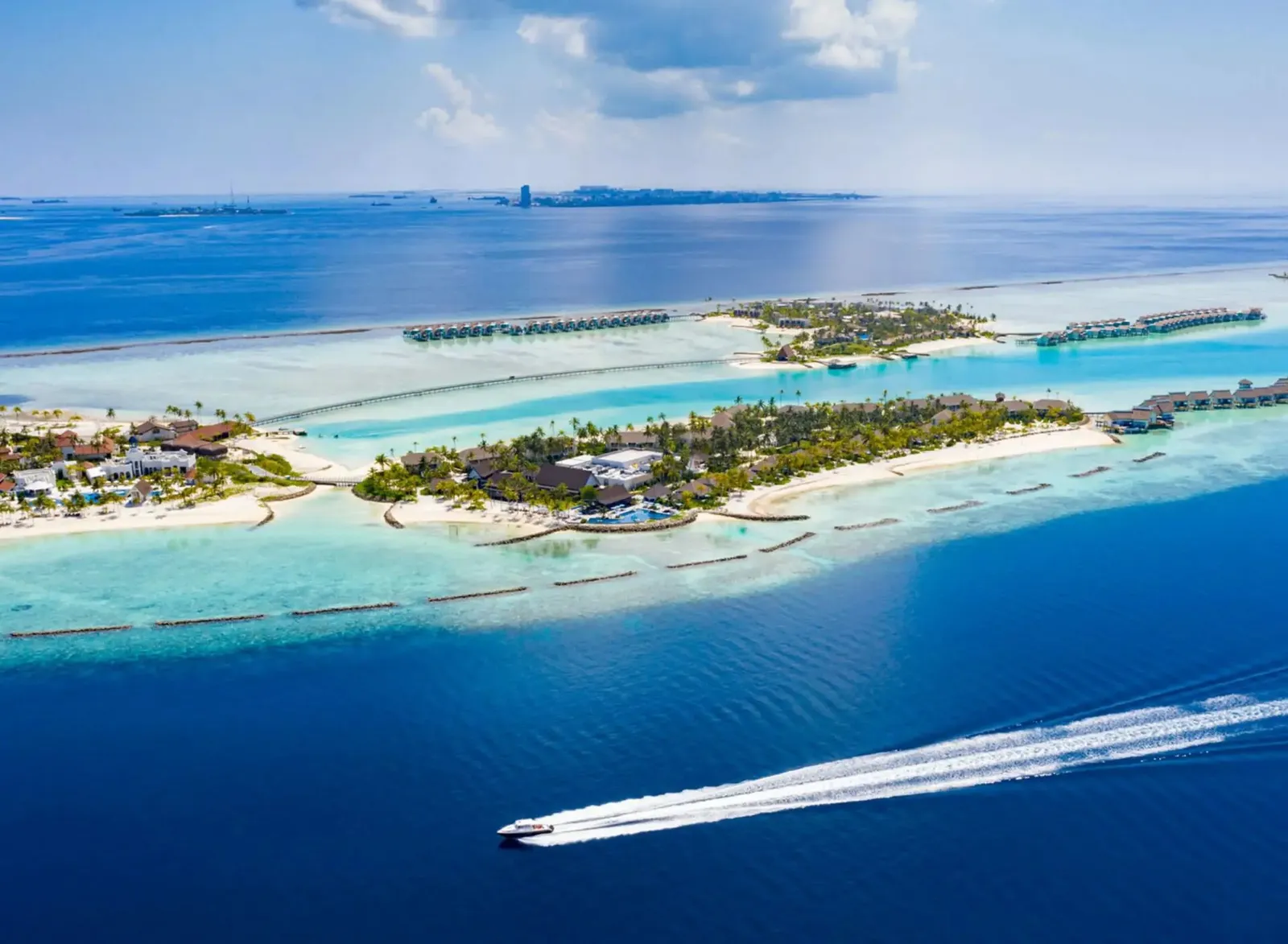 A speedboat’s wake in front of the CROSSROADS islands; on the horizon, the towers of Malé