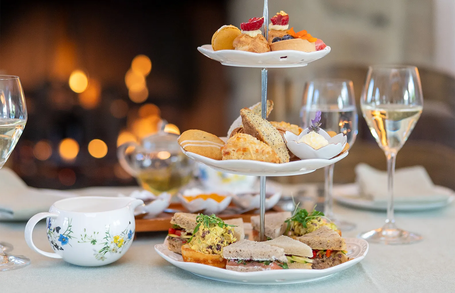 Afternoon tea: a 3-tier stand of sandwiches and pastries, a flowered cup and 2 glasses, by the fire.