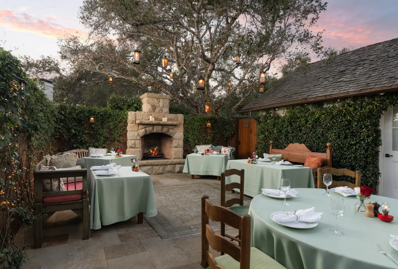 A patio with an outdoor stone fireplace and tables laid beneath an oak at dusk.