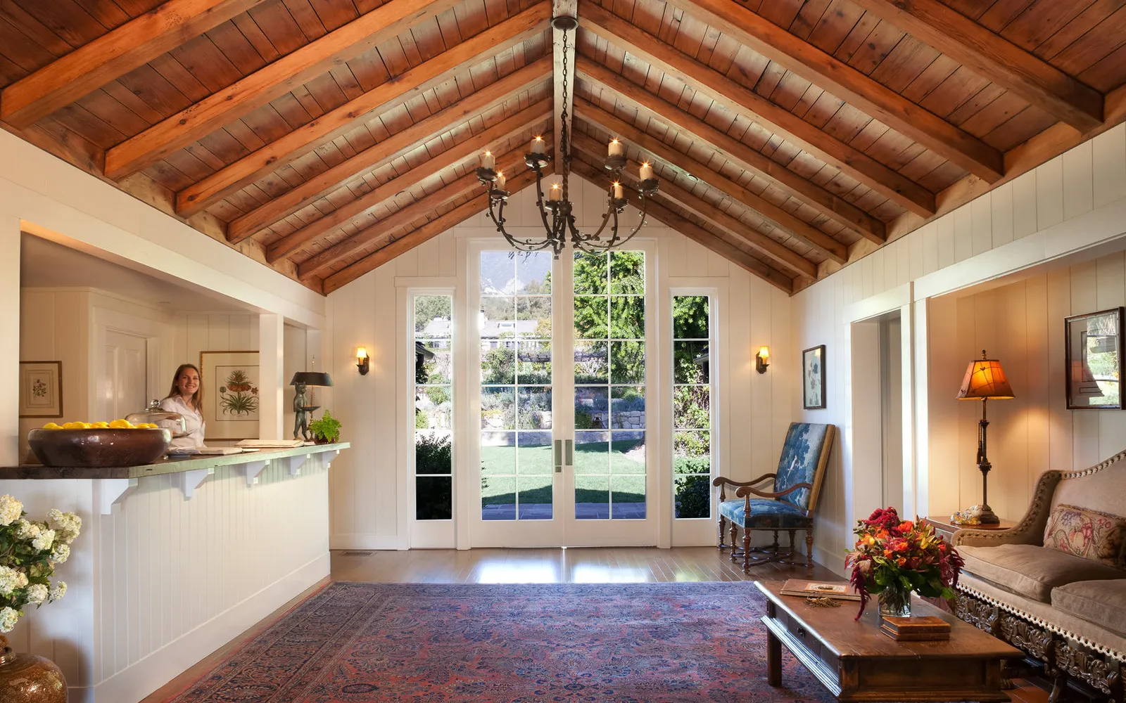 The front desk: a receptionist at the white counter, the trussed timber ceiling, rugs and French doors onto the garden.