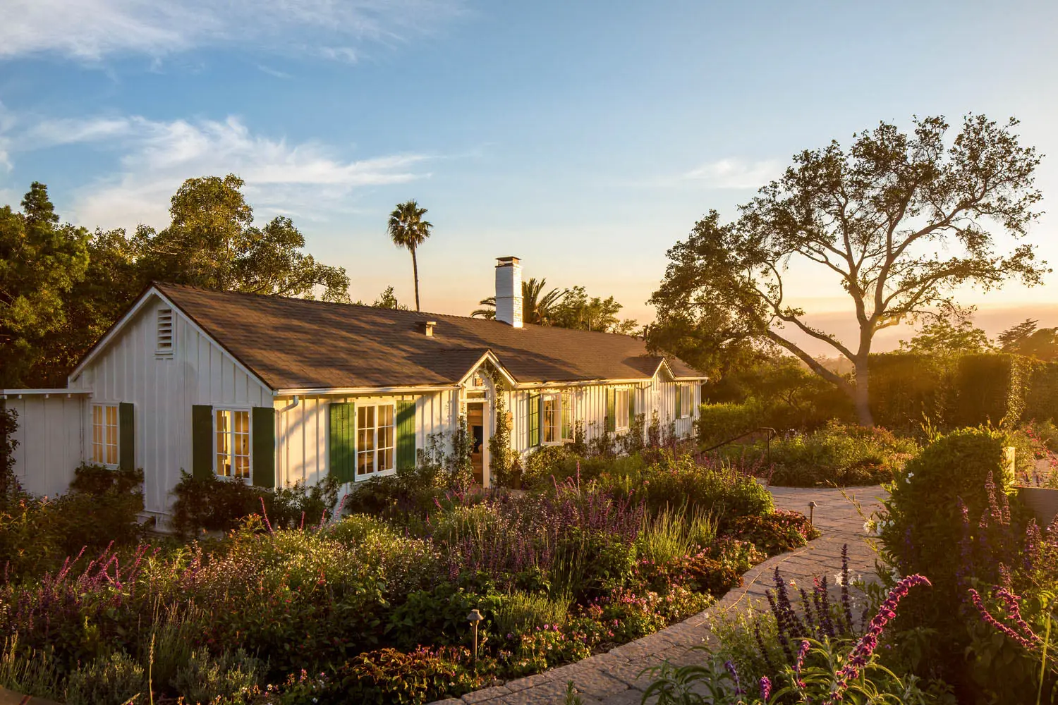 The white Salon & Spa building at sunset, with green shutters, a chimney and a path through flowering shrubs.