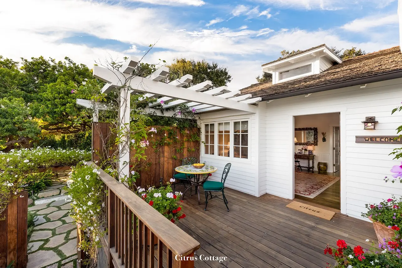 Citrus: the white cottage with its pergola, a wooden deck with a small table and a stone path between flowers.