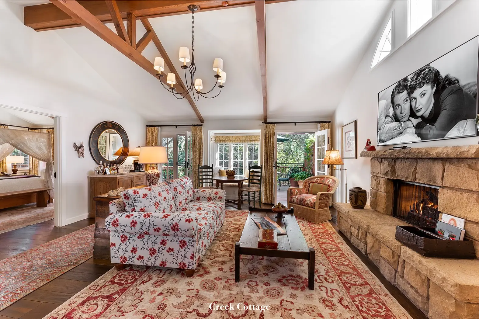 Creek: the living room with its stone fireplace lit, a floral sofa, a trussed ceiling and the bedroom through the door.