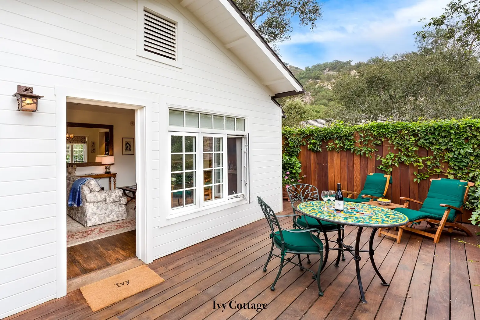 Ivy: the wooden deck with a tiled table, green chairs and the white clapboard cottage.