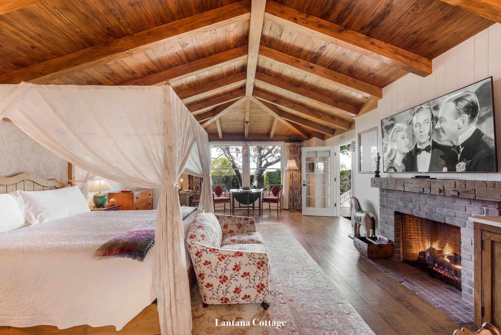Lantana: the canopy bed facing the lit stone fireplace, under a trussed timber ceiling.