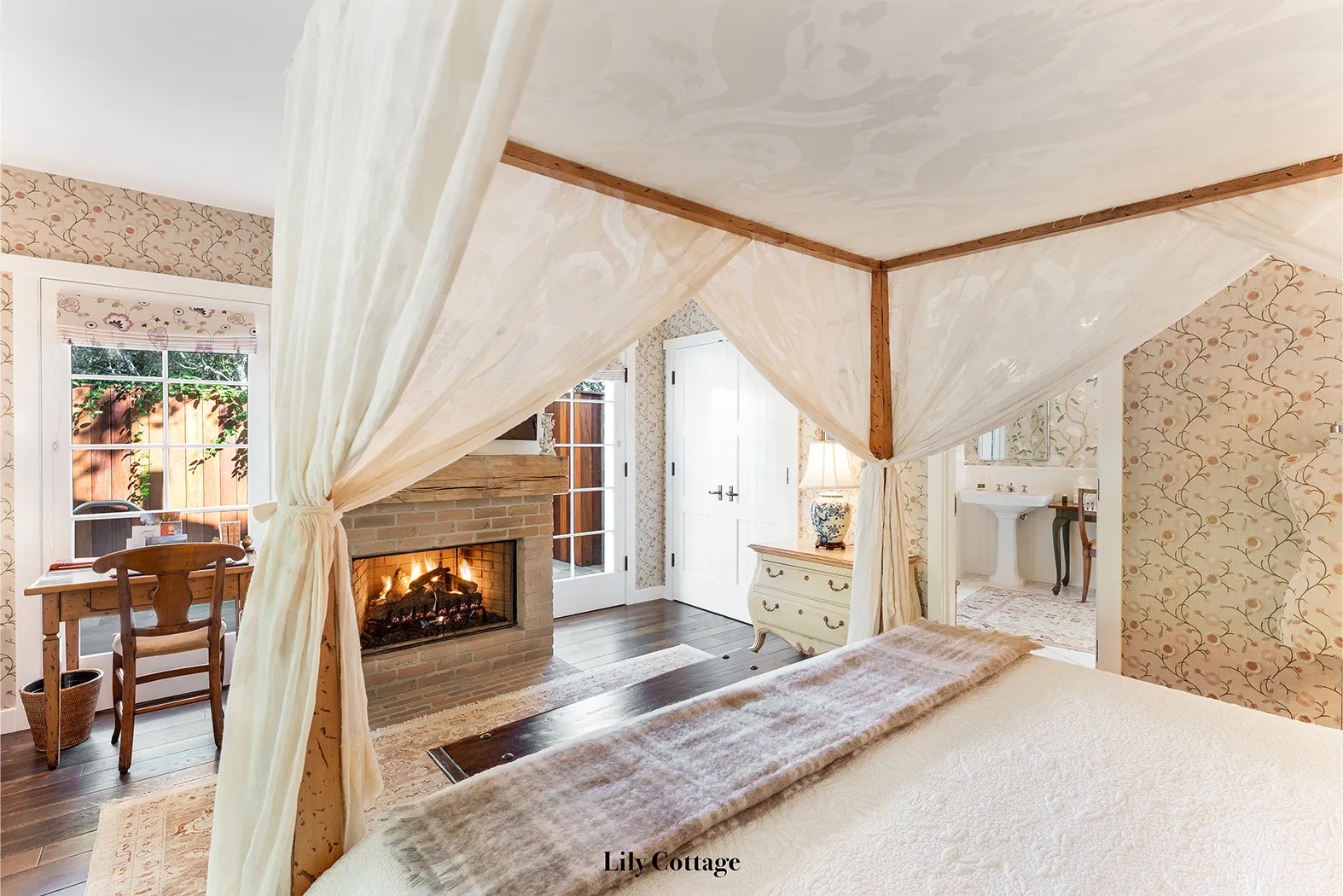 Lily: the canopy bed with pale drapes, facing the lit fireplace.