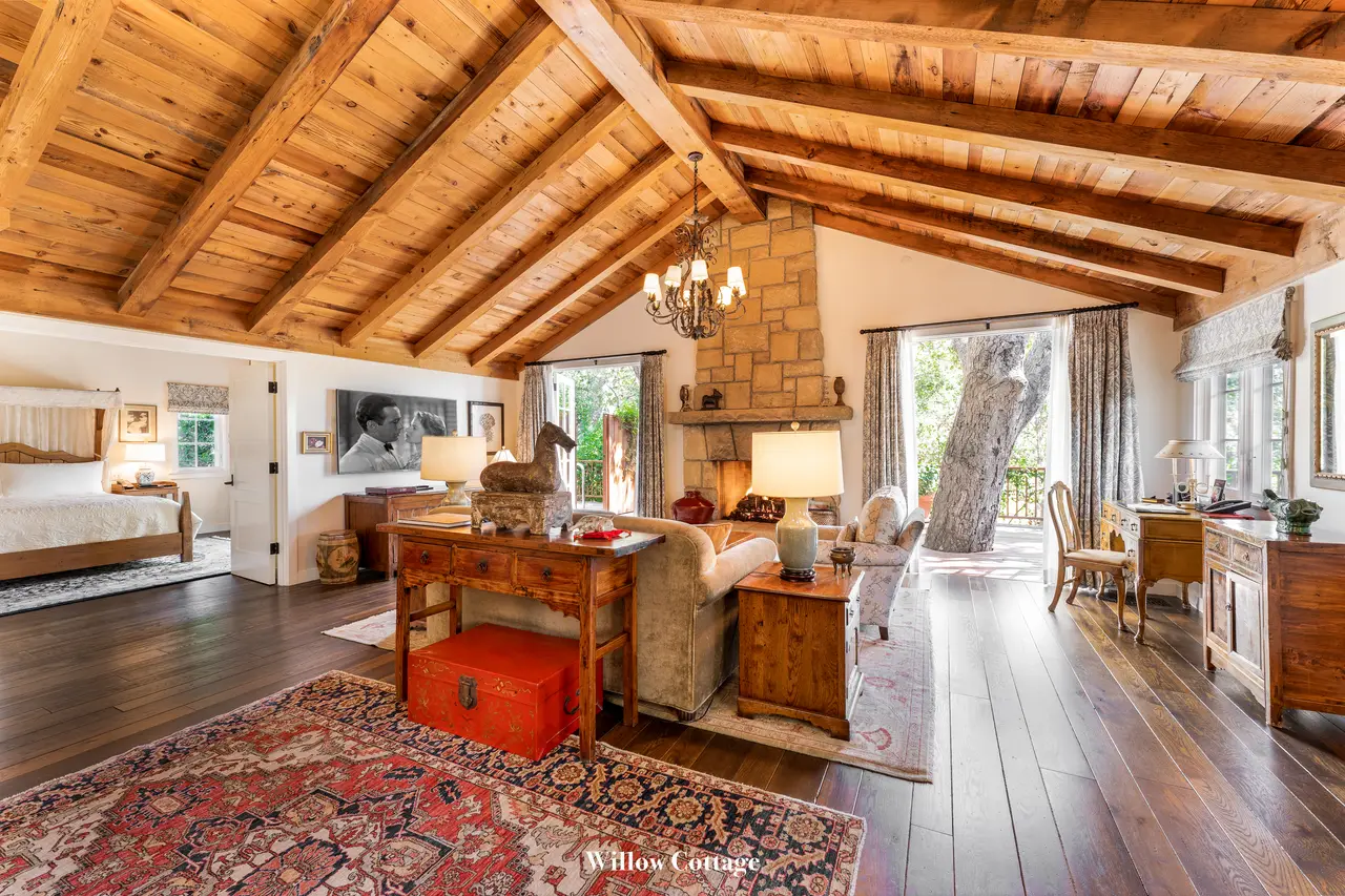 Willow: the living room open to the bedroom, under the timber ceiling, with its stone fireplace and red rugs.