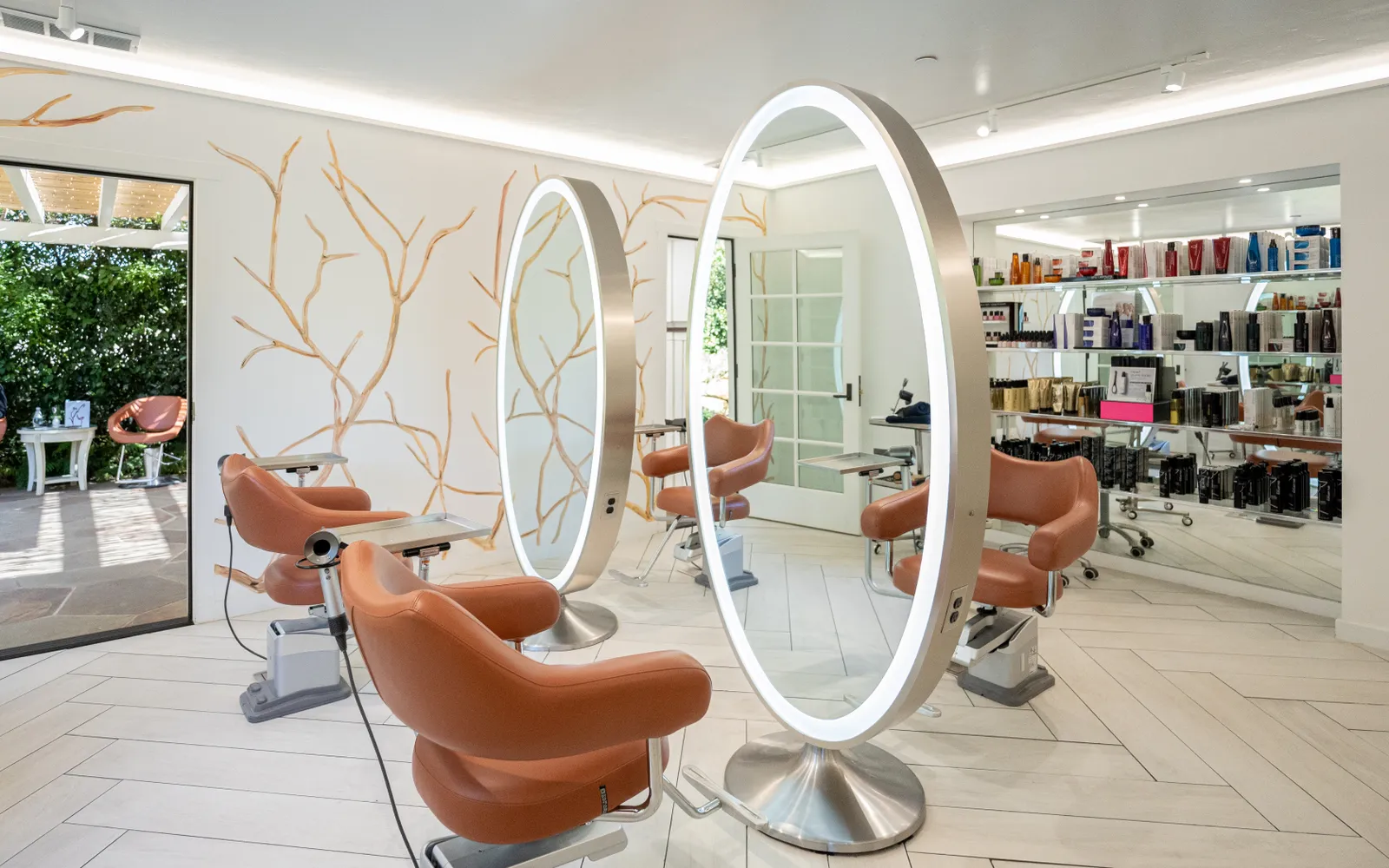The hair salon, with 2 large lit oval mirrors, tan chairs and shelves of products.