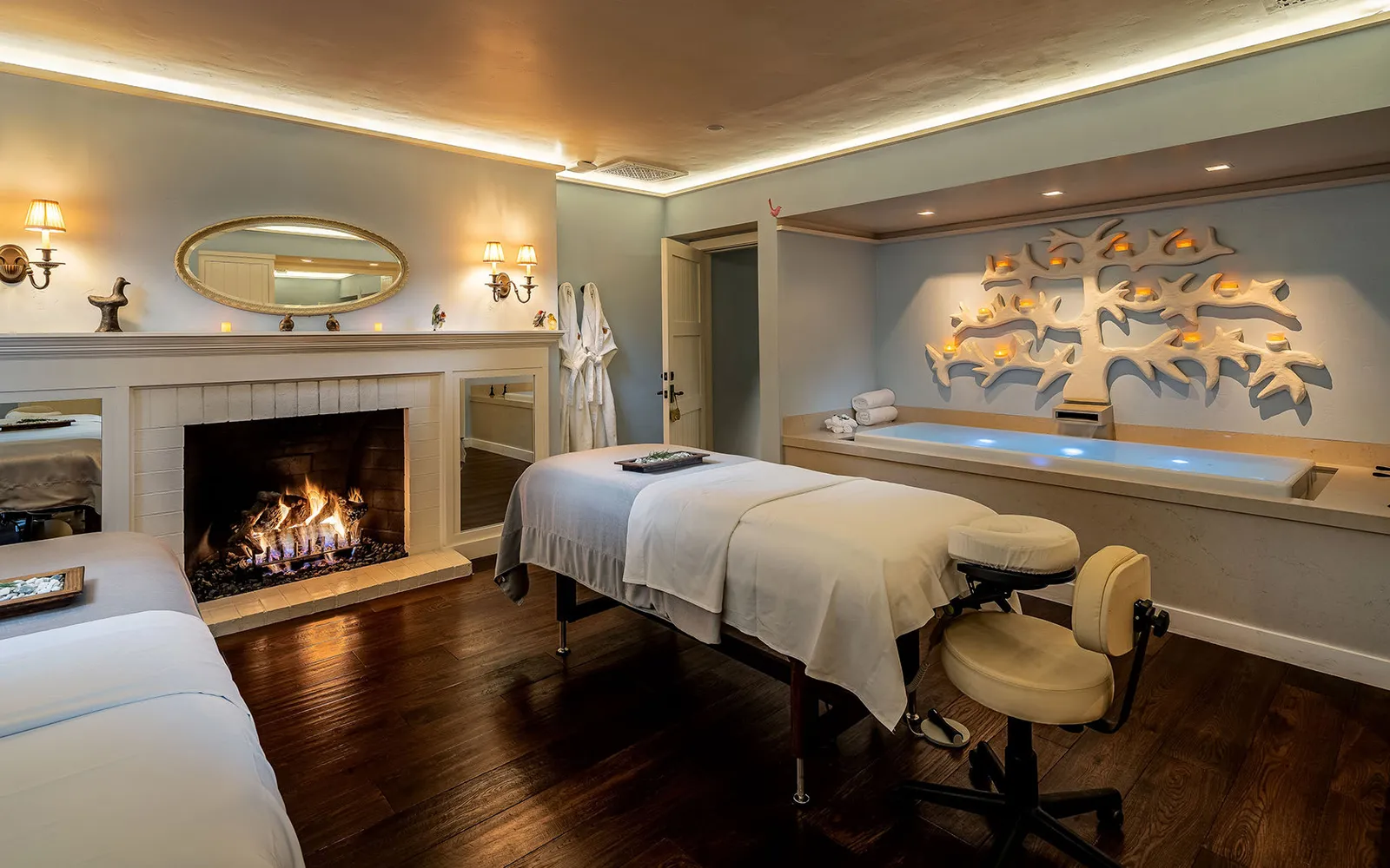 A spa treatment room: the massage table, a lit fireplace and an illuminated tub under a relief of branches.