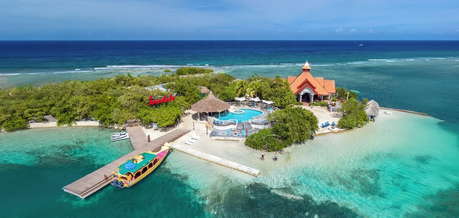 Official photograph, from before the reopening: the private island from above, its pool among the trees, the jetty and the painted boat that runs to and from the resort.