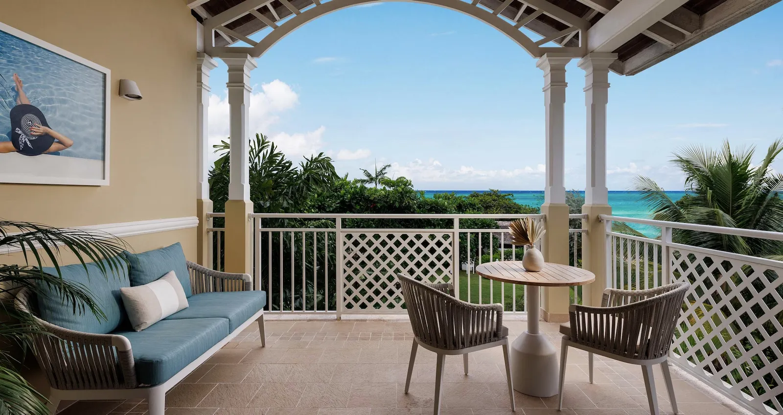 Official image: a veranda with white columns and a lattice rail, a sofa and the sea beyond the garden.
