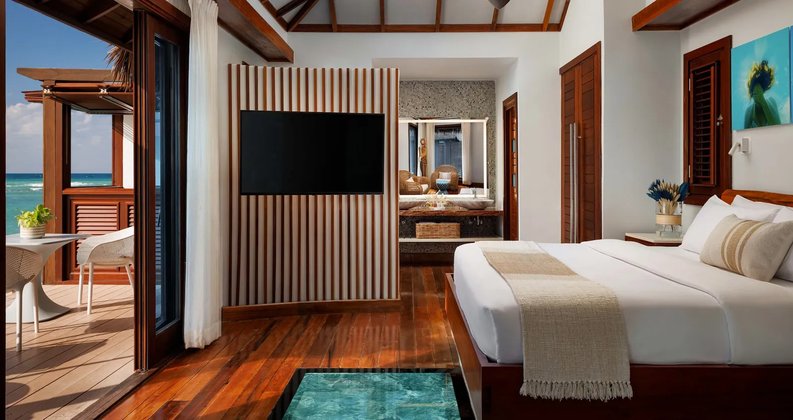 Official photograph, from before the reopening: an over-water bungalow’s bedroom, with its hardwood floor, the glass panel over the sea and the deck.