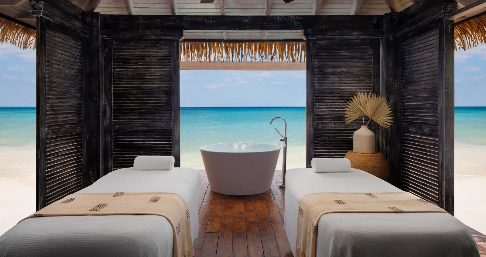 Official image: a treatment cabana open to the sea, with 2 massage beds and a bathtub between dark shutters.
