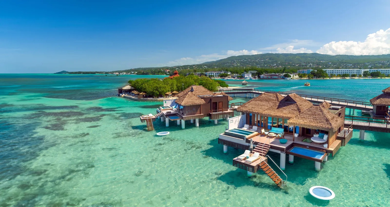 Official photograph, from before the reopening: 2 over-water villas with thatched roofs and infinity pools on their decks, the Montego Bay coast behind.