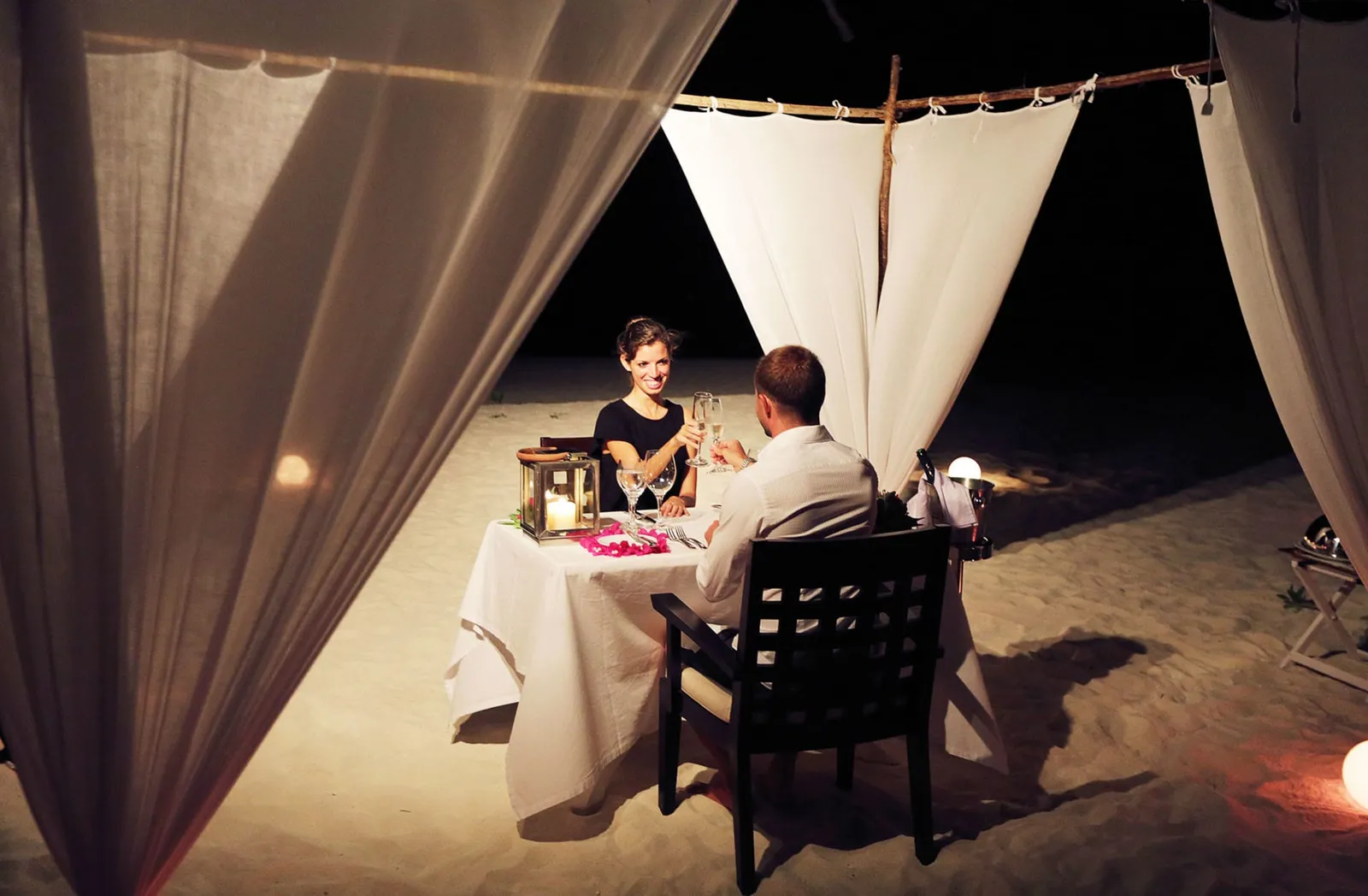 Dinner for 2 on the beach at night, under white drapes, with lanterns on the table.