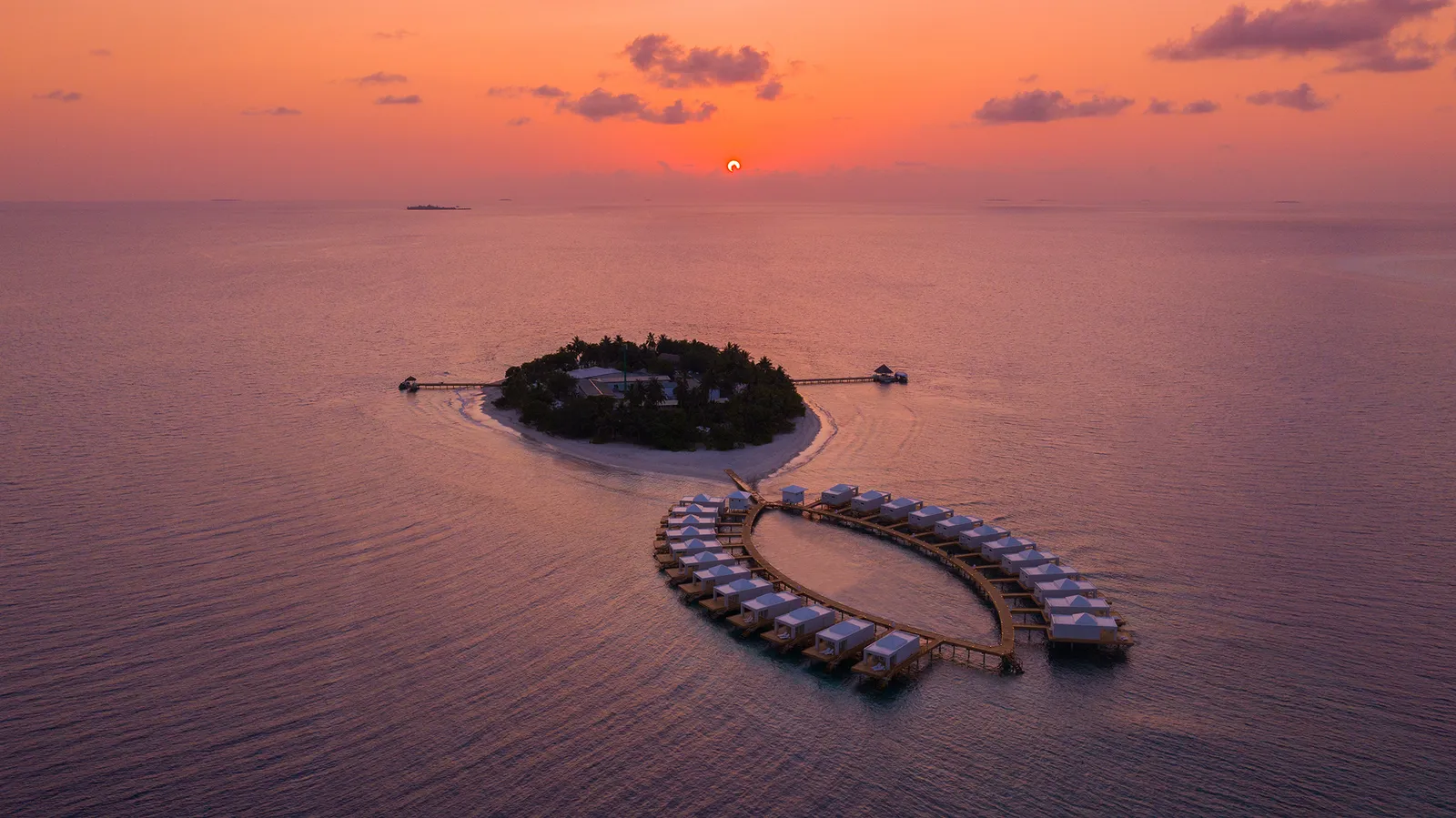The island at sunset from above, the ring of water villas lit and the sea pink all around.