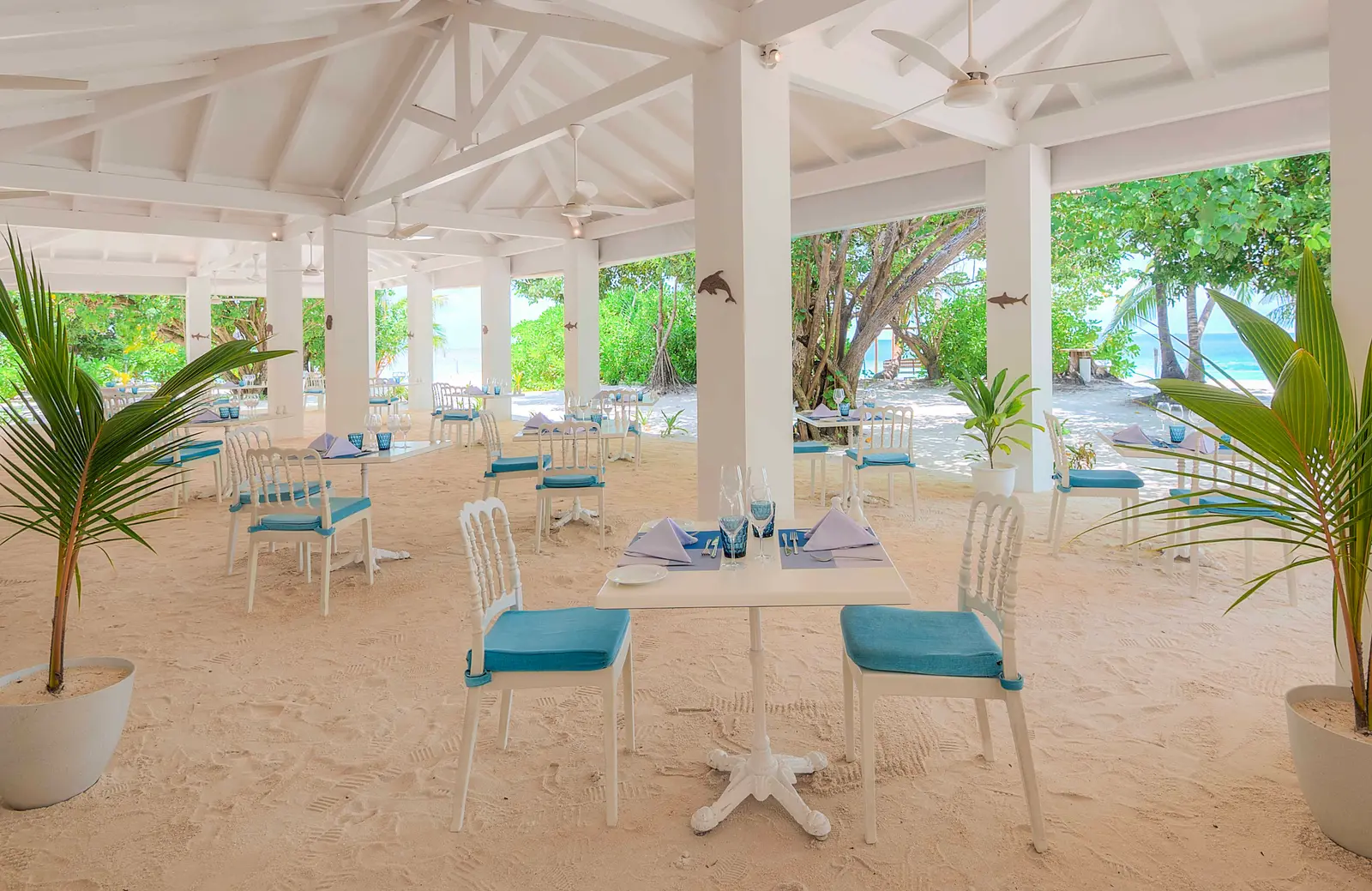 The Island Gallery: white tables and blue chairs on a sand floor, under a white roof open to the palms.