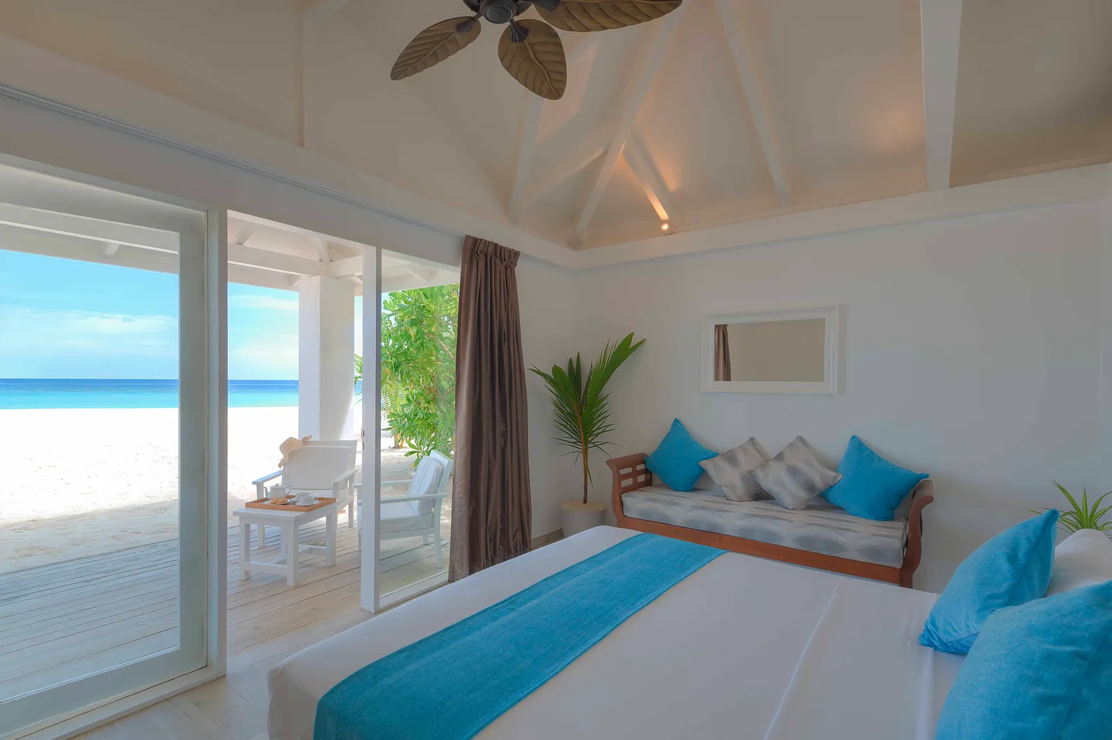 A beach bungalow’s bedroom: the white and blue bed, the daybed and the glass doors open to the sand and the sea.